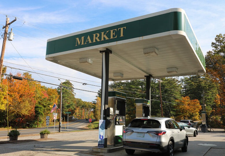 Two gas pumps with an SUV parked next one and autumn weather trees in the background.